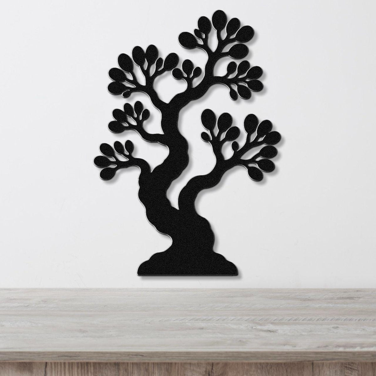 Black metal wall art in the shape of a bonsai tree with detailed branch and leaf cutouts, mounted on a white wall above a wooden surface.
