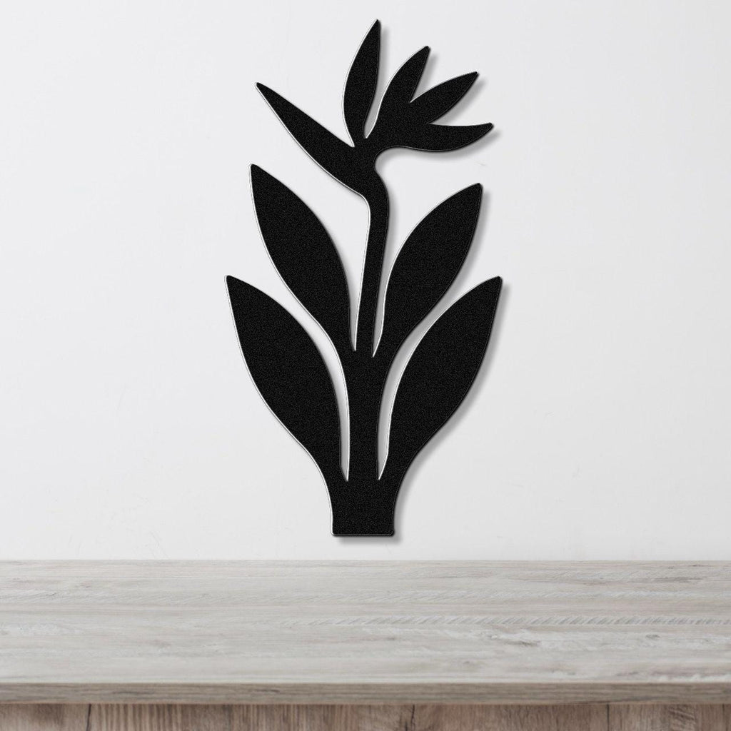 Black metal wall art in the shape of a Bird of Paradise plant with leaf and flower cutouts, mounted on a white wall above a light wood surface.