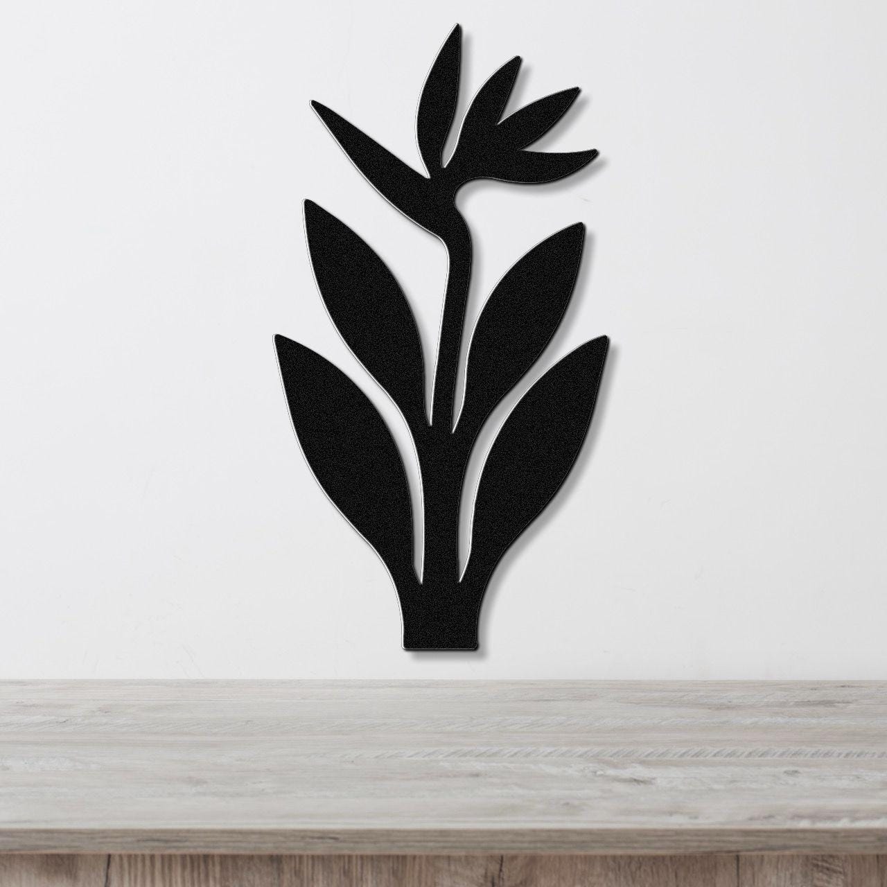 Black metal wall art in the shape of a Bird of Paradise plant with leaf and flower cutouts, mounted on a white wall above a light wood surface.