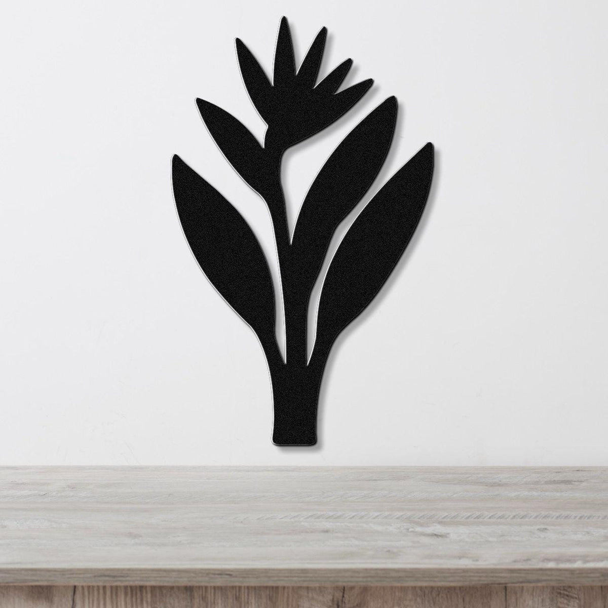 Black metal wall art in the silhouette of a Bird of Paradise plant with cutout leaves and flower, mounted on a white wall above a light wood surface.