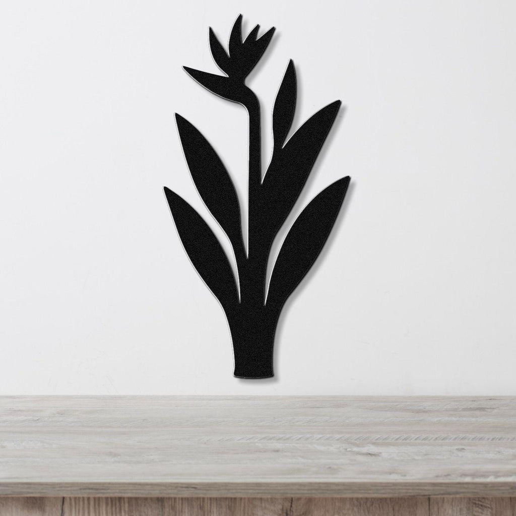 Black metal wall art in the silhouette of a bird of paradise plant with cutout leaves and flower, mounted on a white wall above a light wood surface.