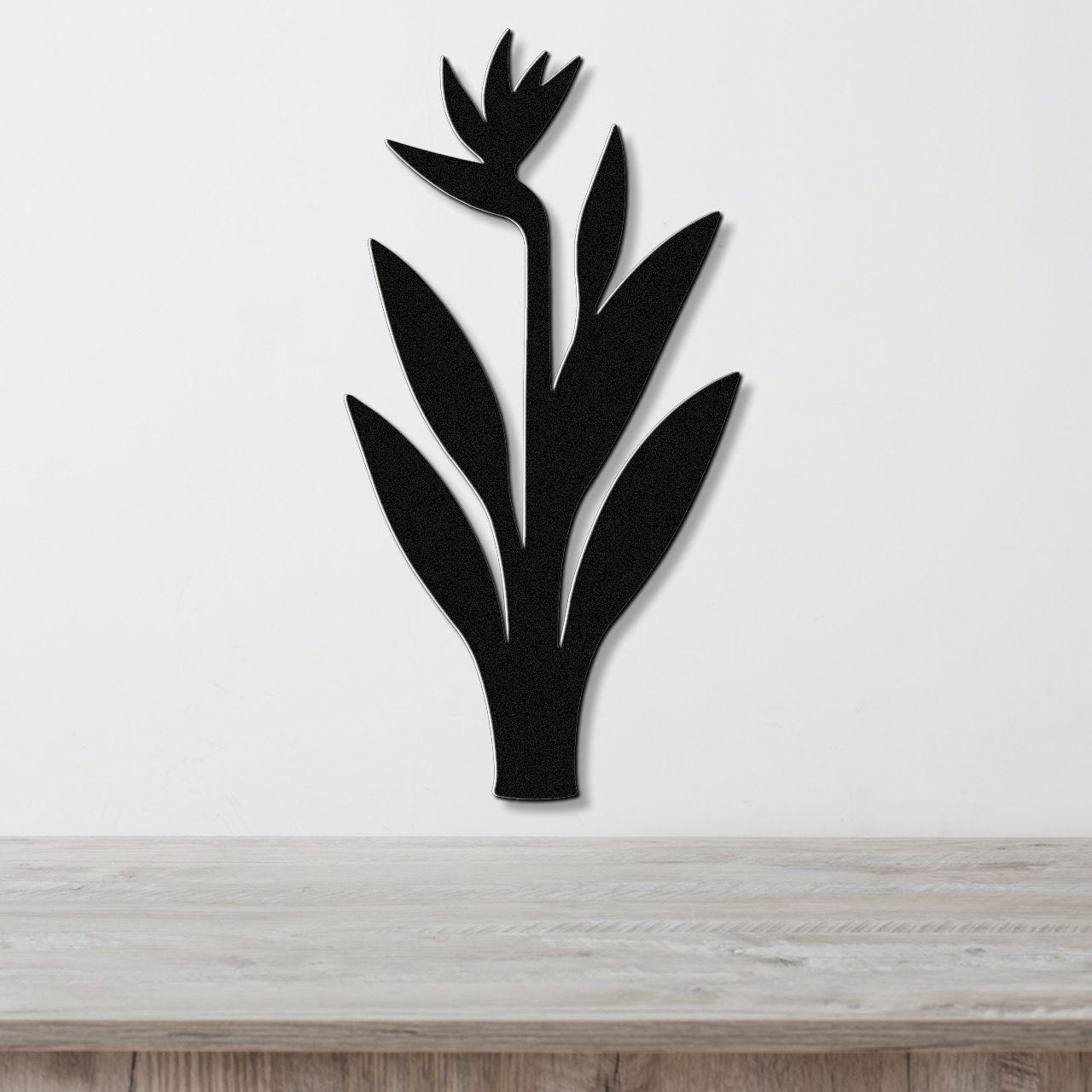 Black metal wall art in the silhouette of a bird of paradise plant with cutout leaves and flower, mounted on a white wall above a light wood surface.