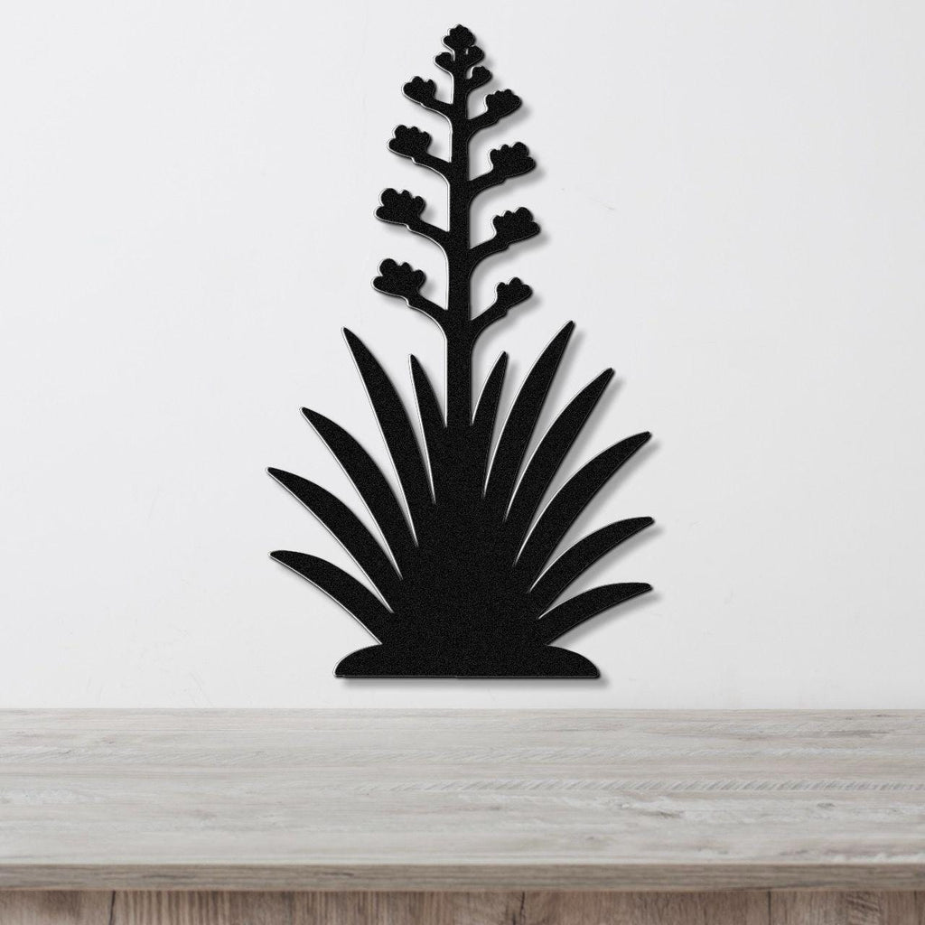 Black metal wall art in the silhouette of a blooming agave plant with a tall flower stalk and spiky leaves, mounted on a white wall above a wooden surface.