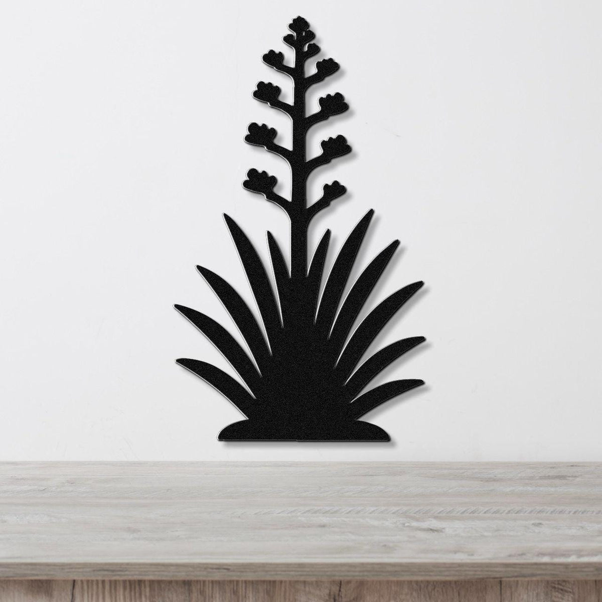 Black metal wall art in the silhouette of a blooming agave plant with a tall flower stalk and spiky leaves, mounted on a white wall above a wooden surface.
