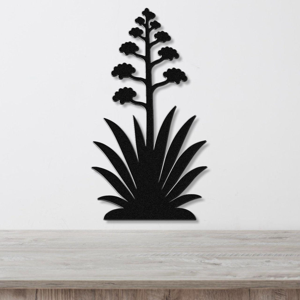 Black metal wall art in the silhouette of a blooming agave plant with a tall flower stalk and spiky leaves, mounted on a white wall above a wooden surface.