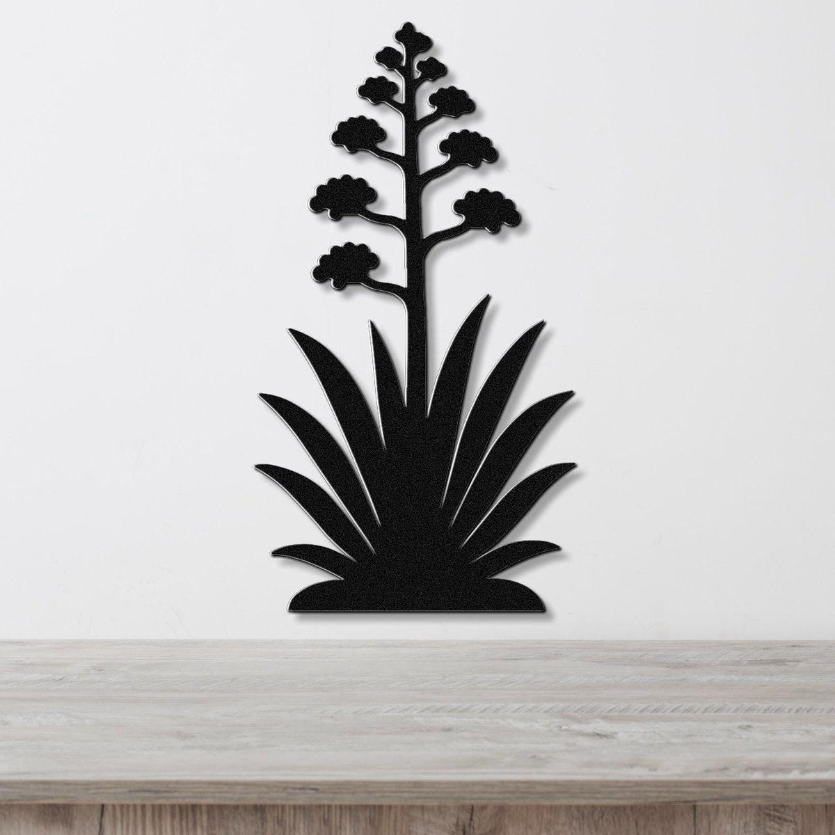Black metal wall art in the silhouette of a blooming agave plant with a tall flower stalk and spiky leaves, mounted on a white wall above a wooden surface.
