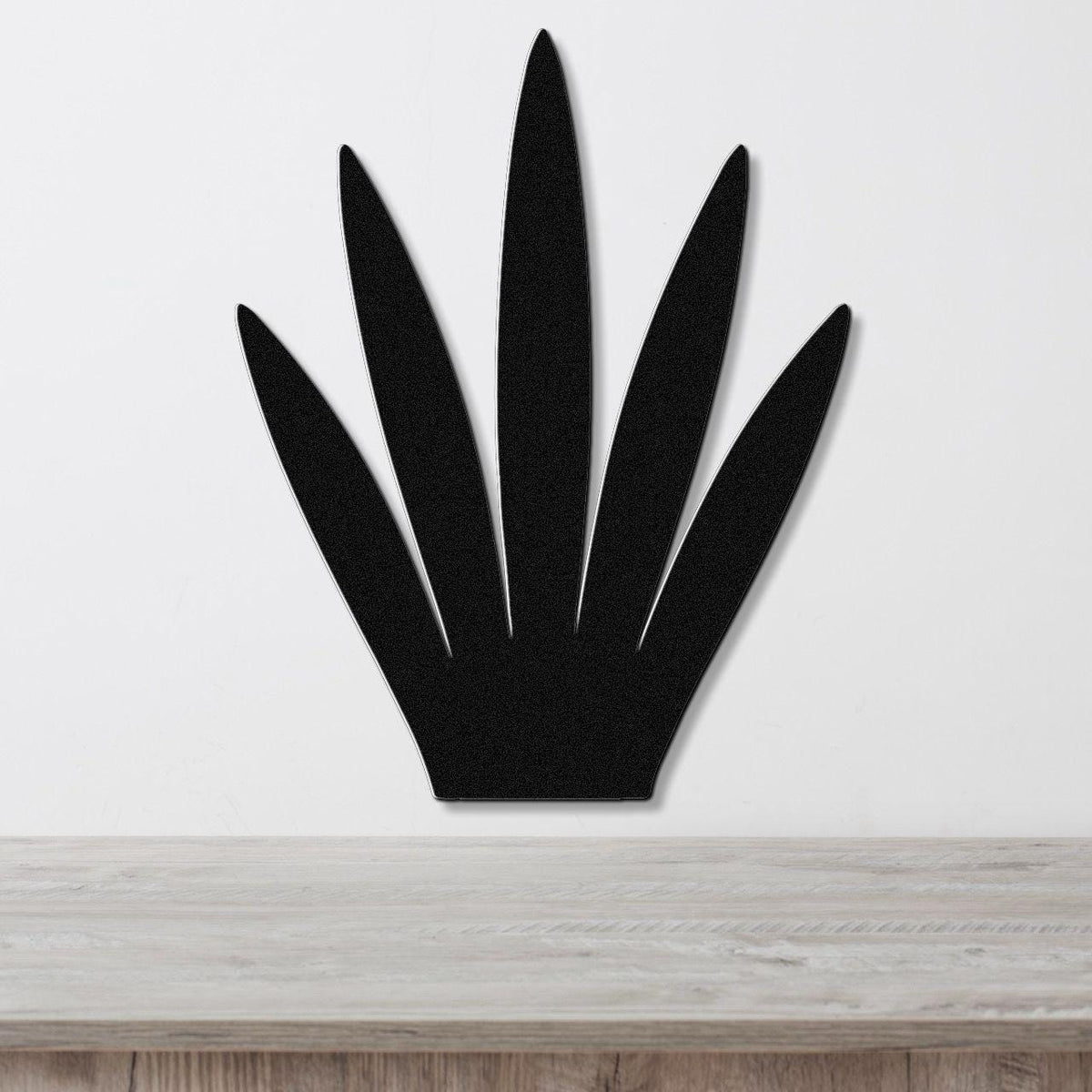 Black metal wall art in the silhouette of a century plant with five pointed leaves, mounted on a white wall above a light wood surface.