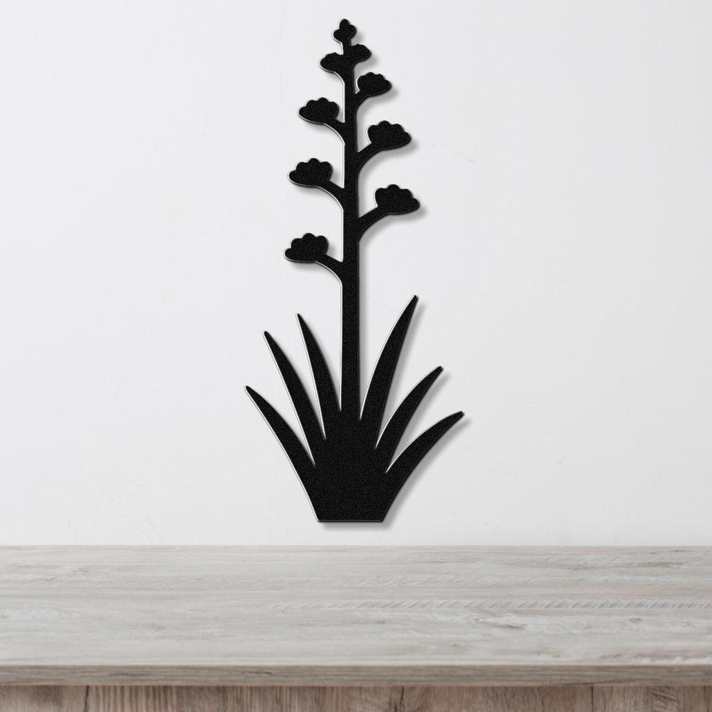 Black metal wall art in the silhouette of a blooming agave plant with a tall stem and spiky leaves, mounted on a white wall above a wooden surface.