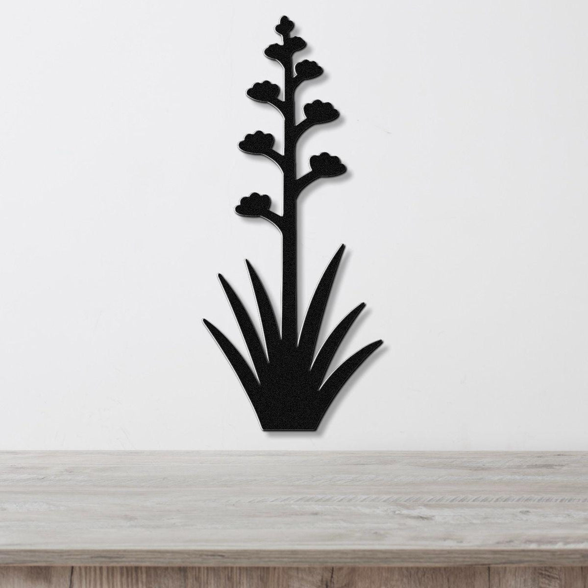 Black metal wall art in the silhouette of a blooming agave plant with a tall stem and spiky leaves, mounted on a white wall above a wooden surface.