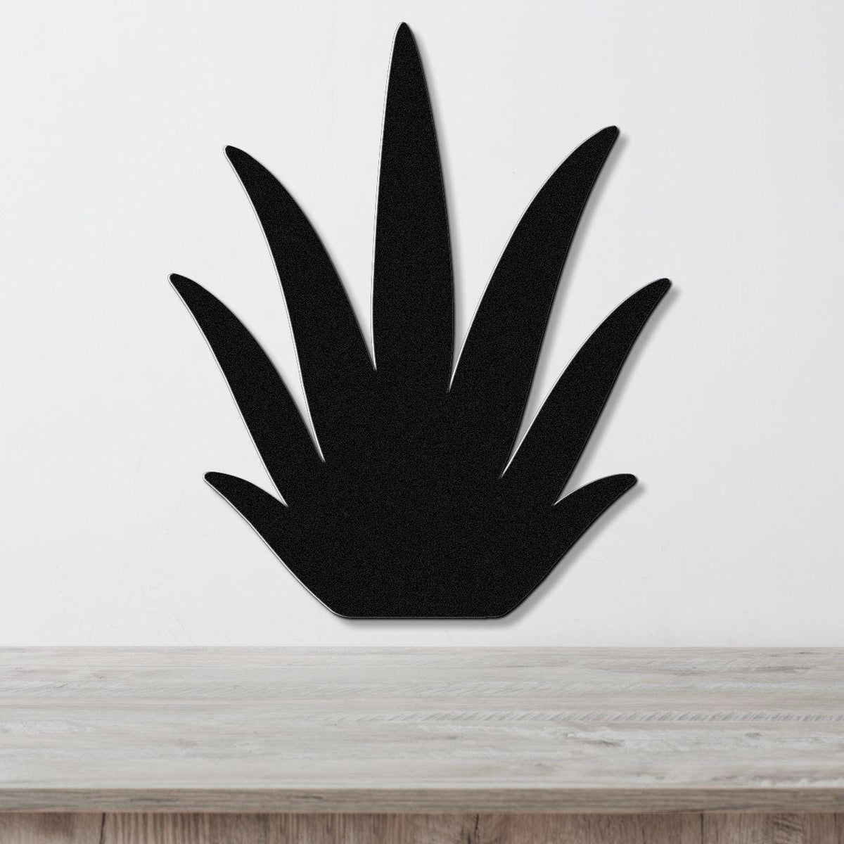 Black metal agave wall art with pointed leaf cutouts, mounted on a white wall above a light wood surface.