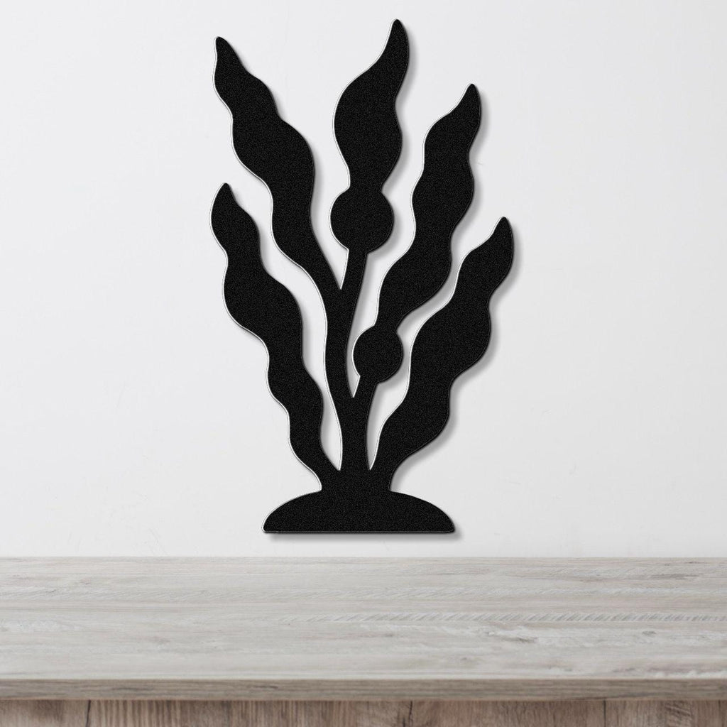 Black metal wall art in a kelp and seaweed silhouette design with internal cutouts, mounted on a white wall above a light wood surface.