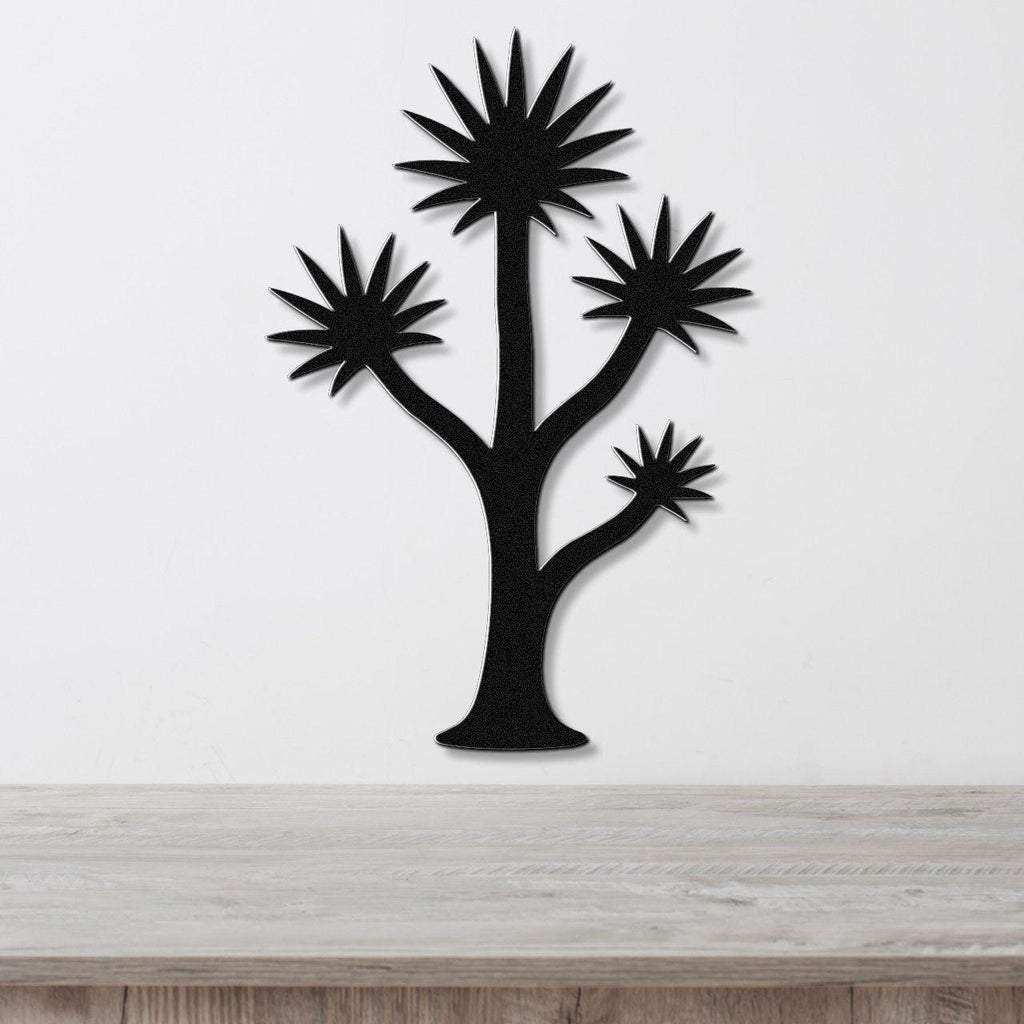 Black metal wall art in a yucca tree silhouette with spiky starburst leaves, mounted on a white wall above a light wood surface.