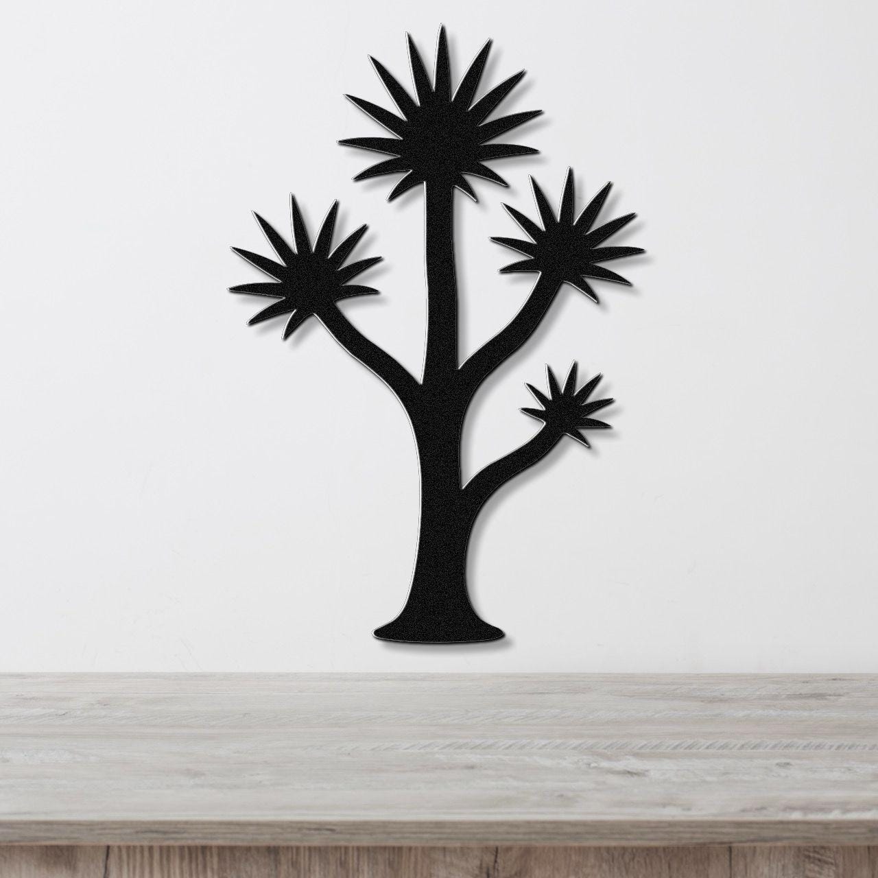 Black metal wall art in a yucca tree silhouette with spiky starburst leaves, mounted on a white wall above a light wood surface.