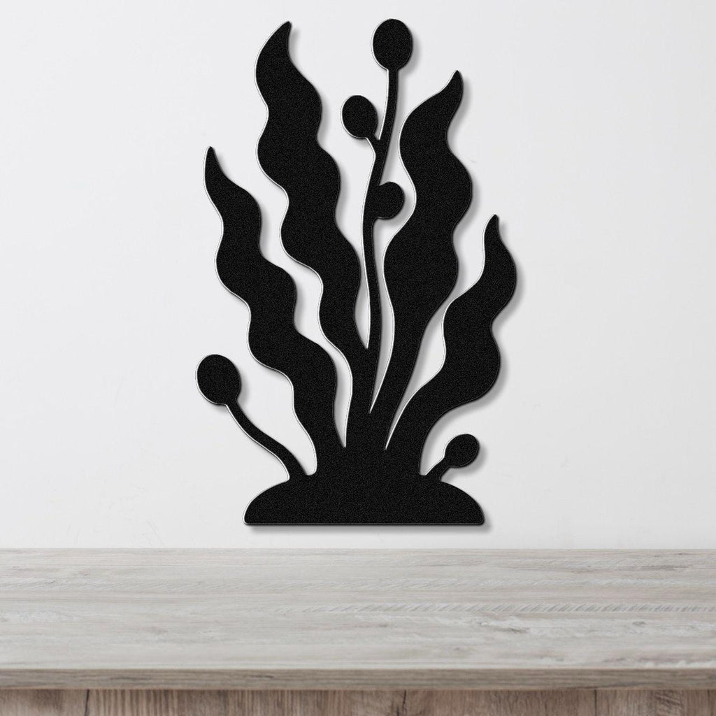 Black metal wall art cutout of kelp and seaweed with wavy fronds and bulbous tips, mounted on a white wall above a light wood surface.