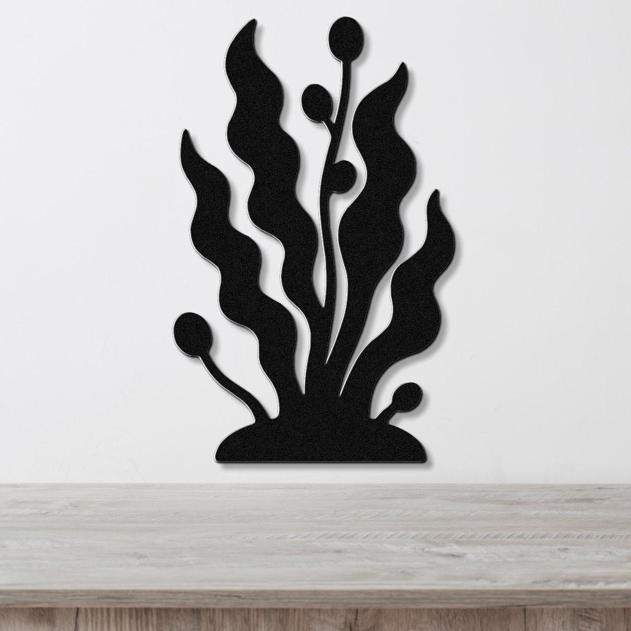Black metal wall art cutout of kelp and seaweed with wavy fronds and bulbous tips, mounted on a white wall above a light wood surface.
