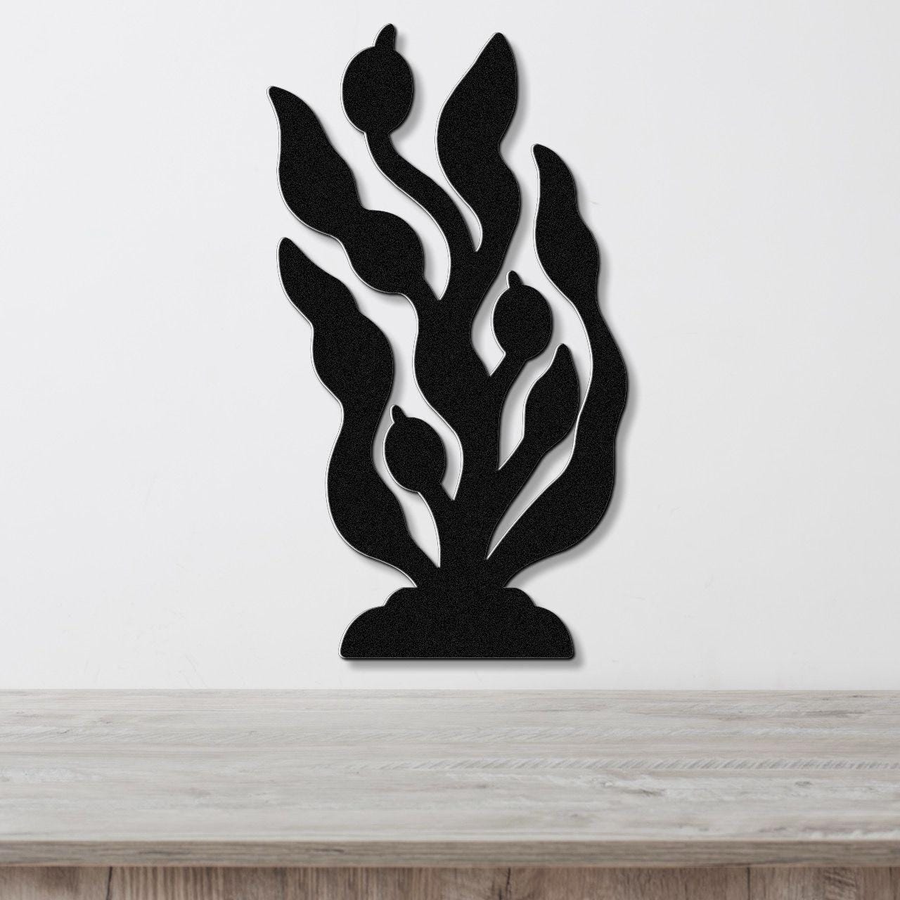 Black metal wall art cutout of kelp and seaweed with flowing fronds and bulbous tips, mounted on a white wall above a light wood surface.