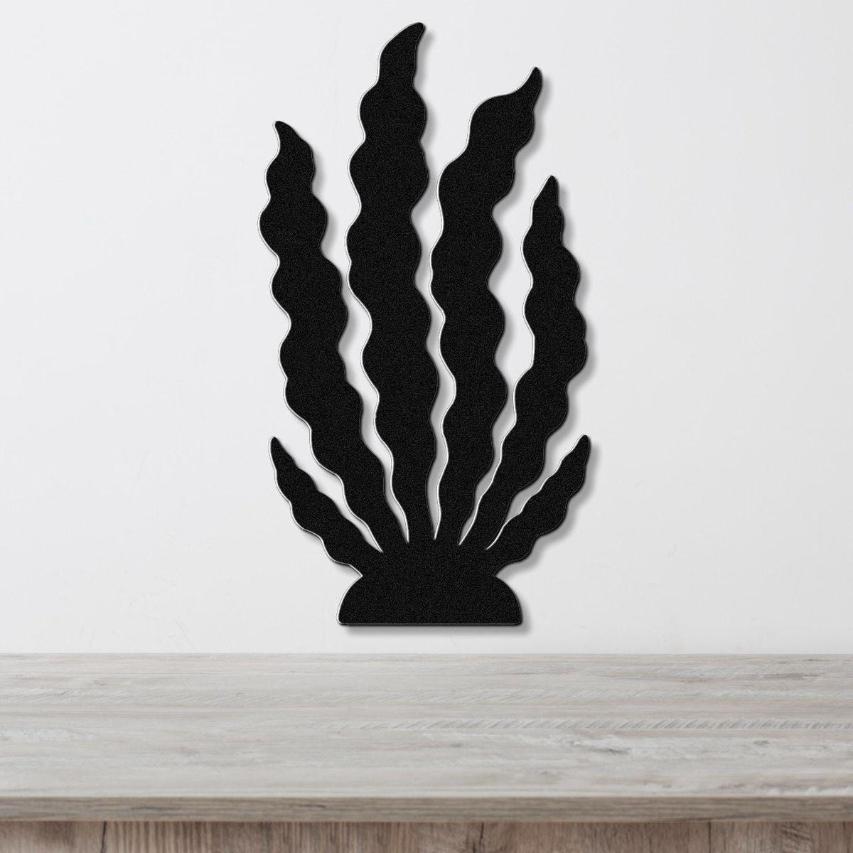 Black metal wall art in a kelp or seaweed silhouette design with wavy fronds, mounted on a white wall above a light wood surface.