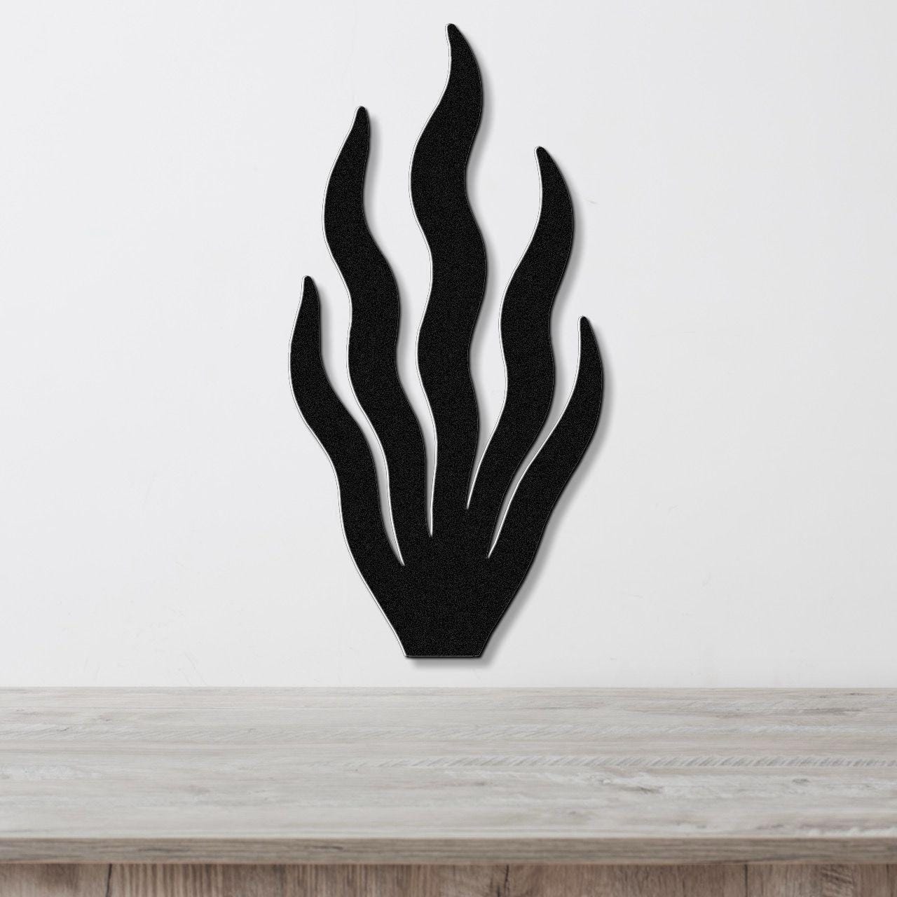 Black metal wall art in a kelp or seaweed silhouette with flowing, wavy fronds, mounted on a white wall above a light wood surface.