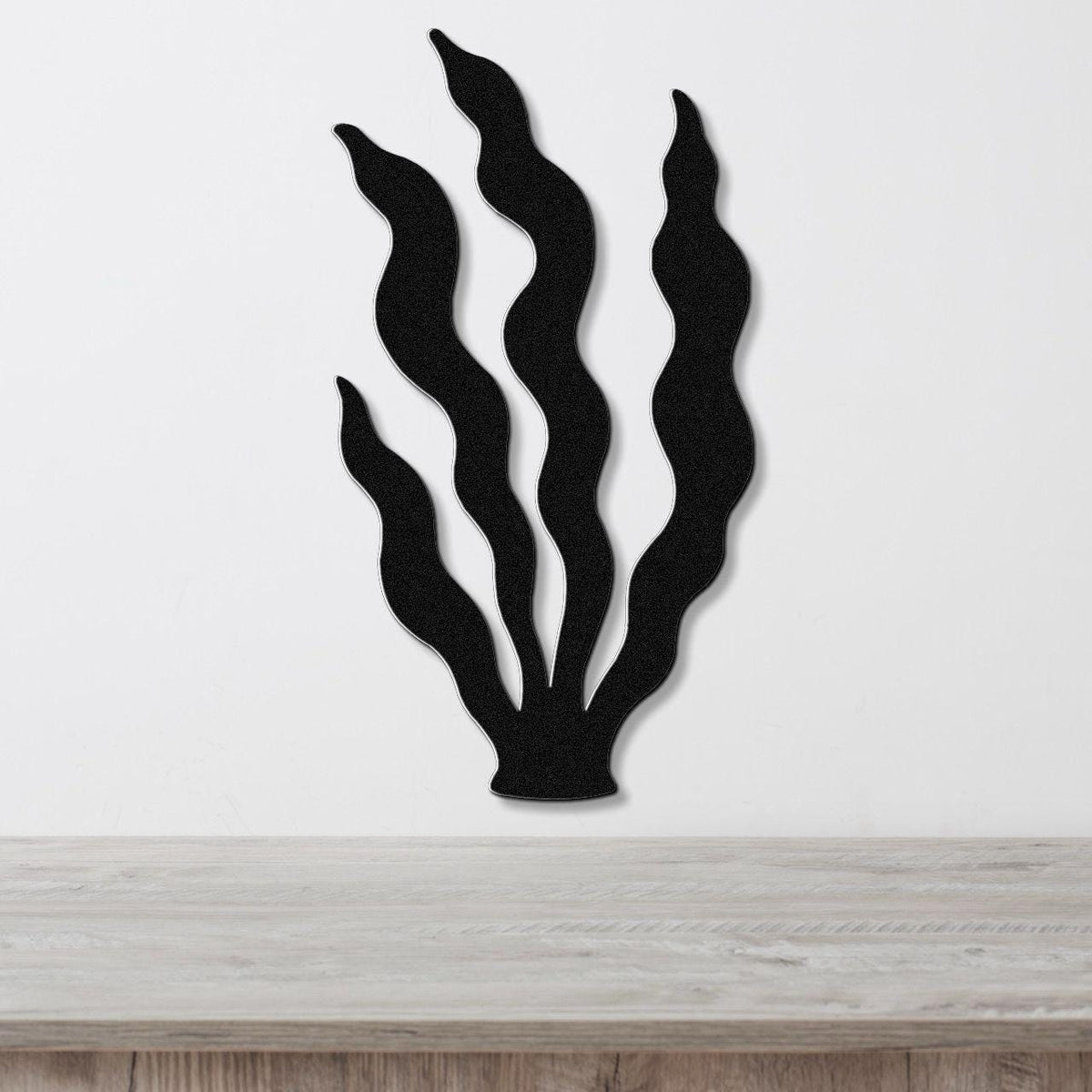 Black metal wall art in a kelp silhouette design with wavy fronds, mounted on a white wall above a light wood surface.