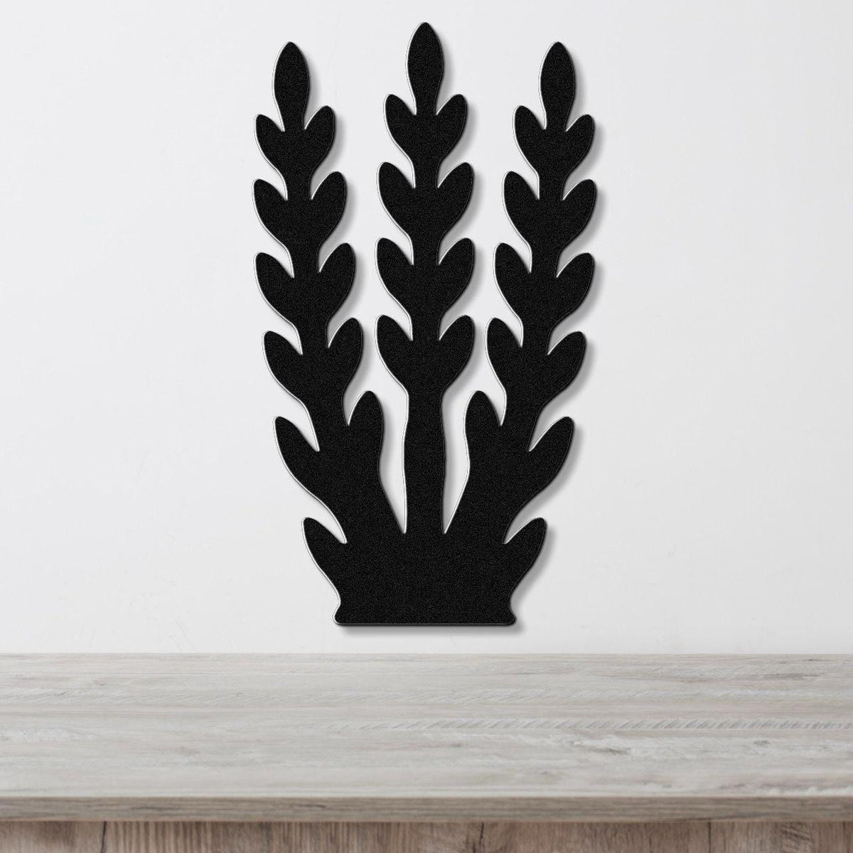 Black metal wall art in a kelp or seaweed silhouette design with layered leaf cutouts, mounted on a white wall above a light wood surface.