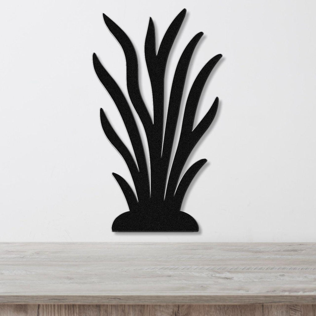 Black metal wall art in a kelp or seaweed silhouette design with layered leaf cutouts, mounted on a white wall above a light wood surface.