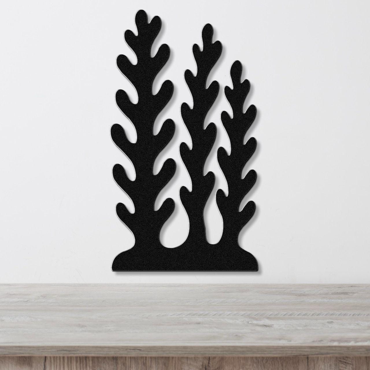 Black metal wall art with a kelp and seaweed silhouette design, featuring three tall, branching cutouts, mounted on a white wall above a wooden surface.