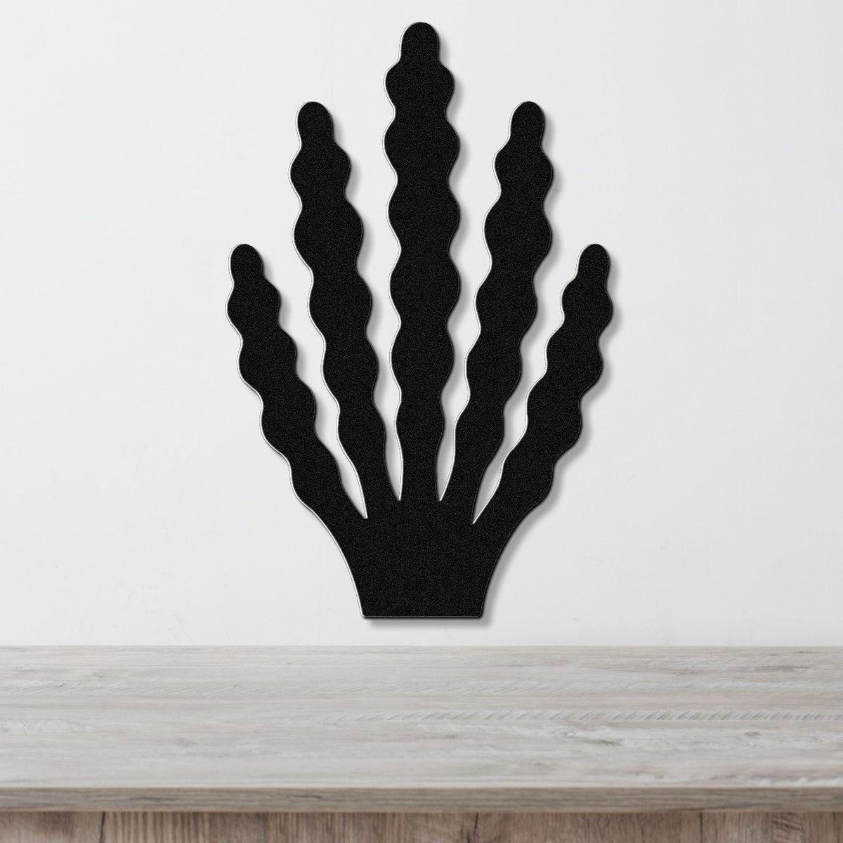 Black metal wall art in a kelp or seaweed silhouette with wavy fronds, mounted on a white wall above a light wood surface.