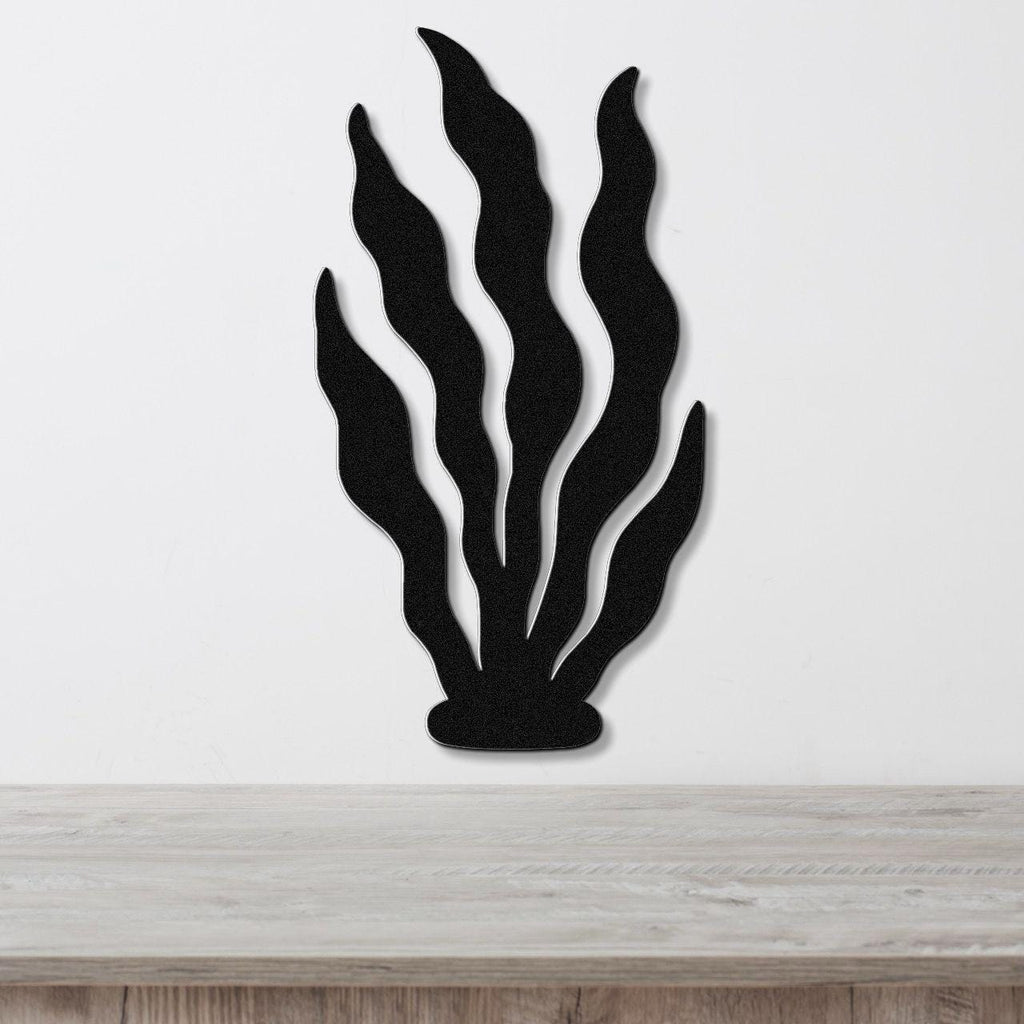 Black metal wall art in a kelp or seaweed silhouette with wavy leaf cutouts, mounted on a white wall above a light wood surface.