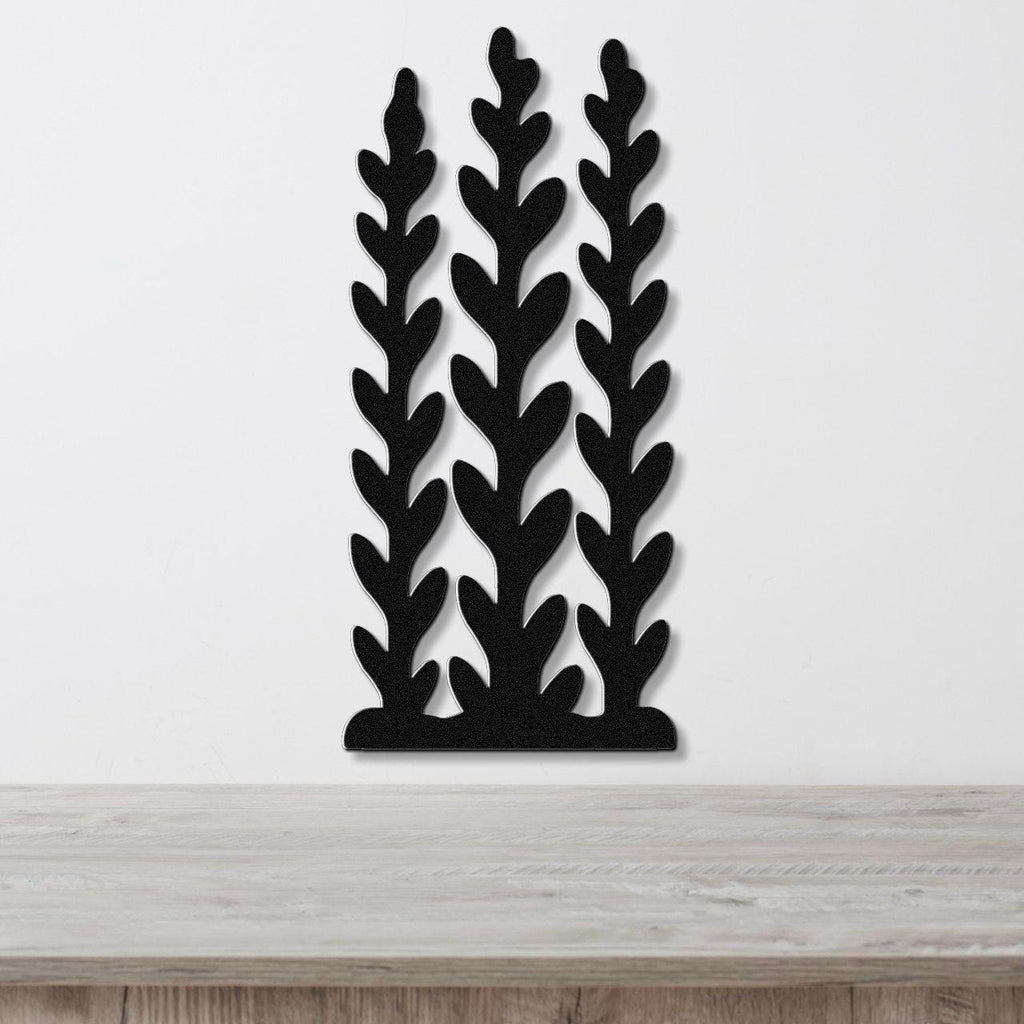 Black metal wall art with a kelp and seaweed silhouette design, featuring three vertical fronds with leaf-like cutouts, mounted on a white wall above a wooden surface.