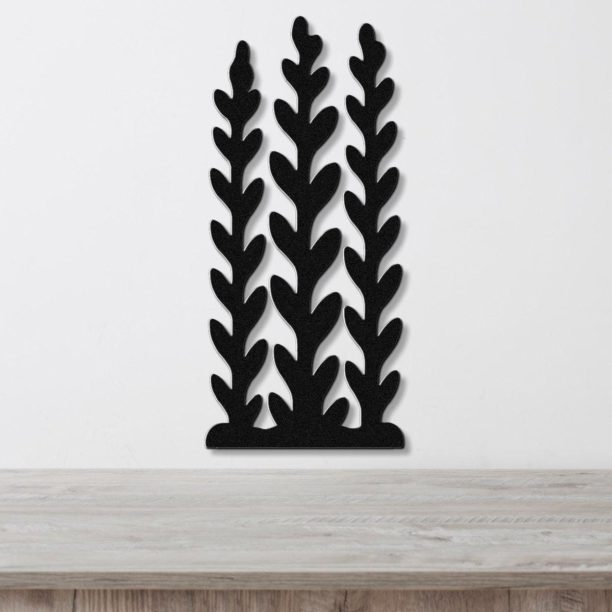 Black metal wall art with a kelp and seaweed silhouette design, featuring three vertical fronds with leaf-like cutouts, mounted on a white wall above a wooden surface.