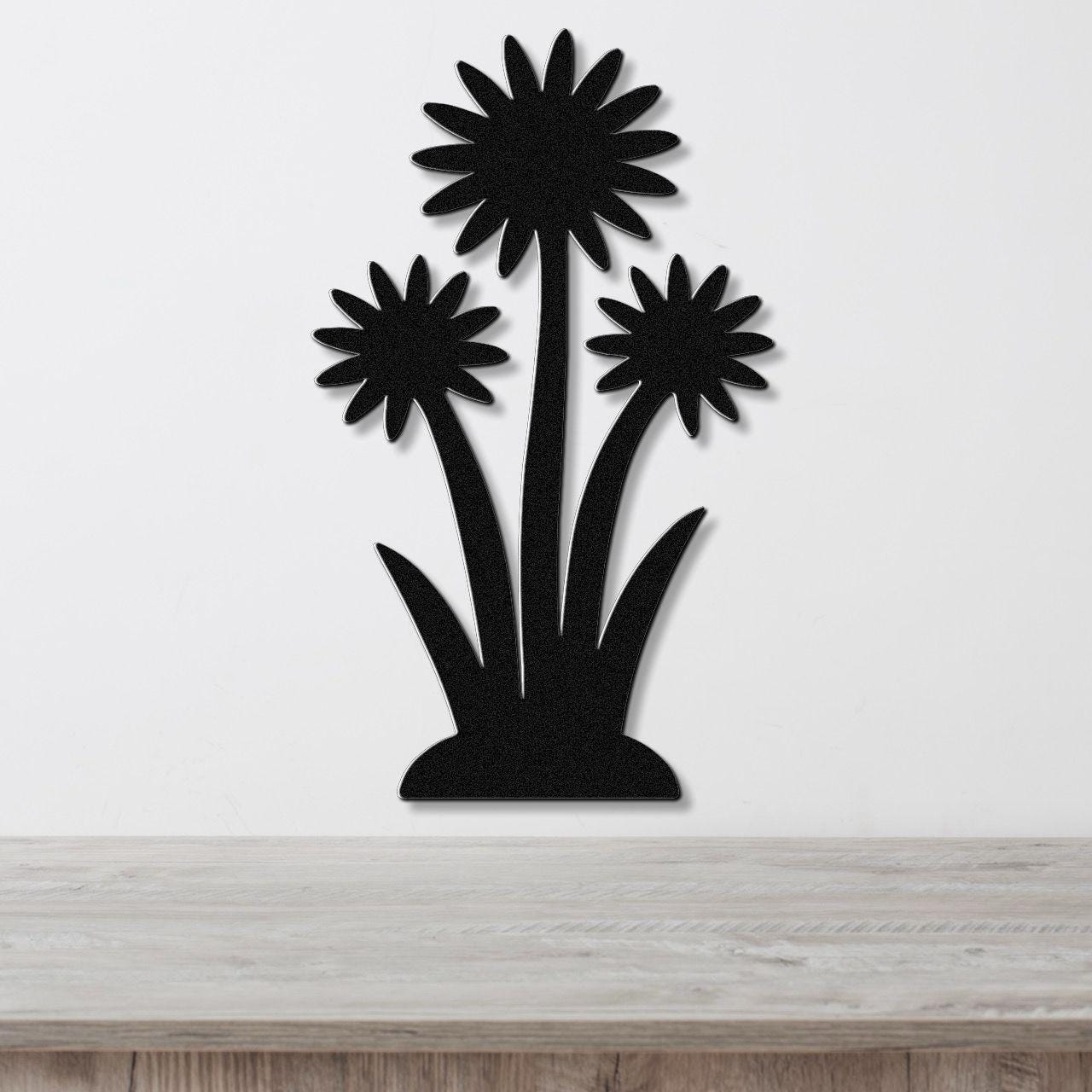 Black metal wall art featuring a silhouette of three wildflowers with long stems and pointed leaves, mounted on a white wall above a light wood surface.