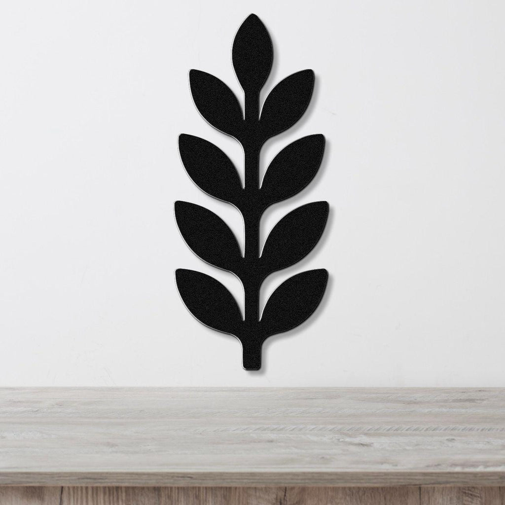 Black metal wall art in a leafy sprig silhouette with cutout details, mounted on a white wall above a light wood surface.