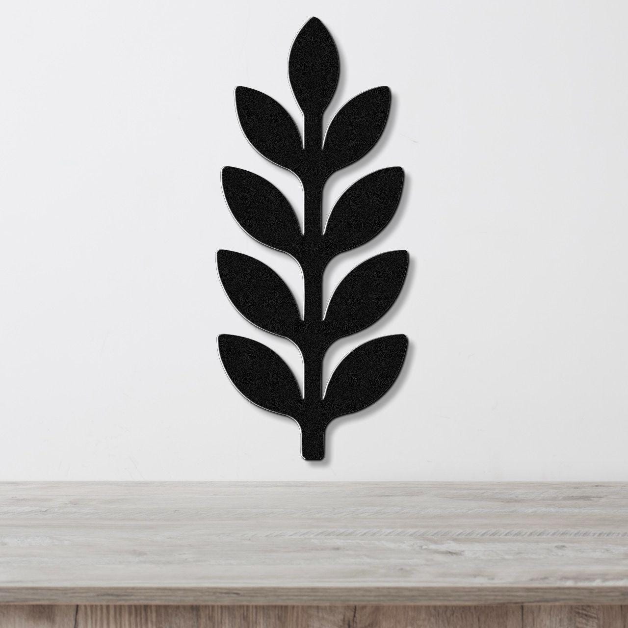 Black metal wall art in a leafy sprig silhouette with cutout details, mounted on a white wall above a light wood surface.