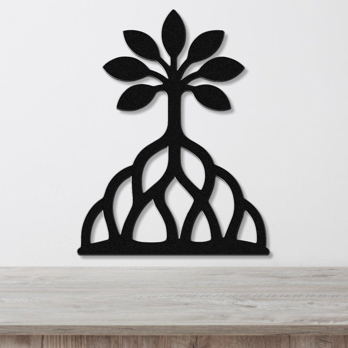 Black metal wall art in the shape of a tree with leafy top and exposed root system, mounted on a white wall above a wooden surface.