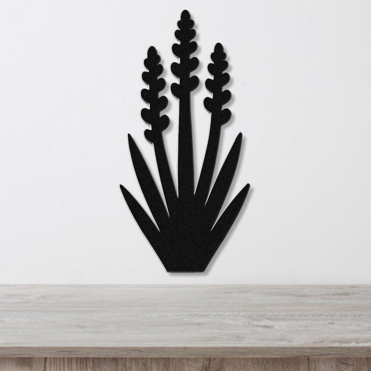 Black metal wall art in a yucca plant silhouette with three tall flower spikes and spiky leaves, mounted on a white wall above a light wood surface.