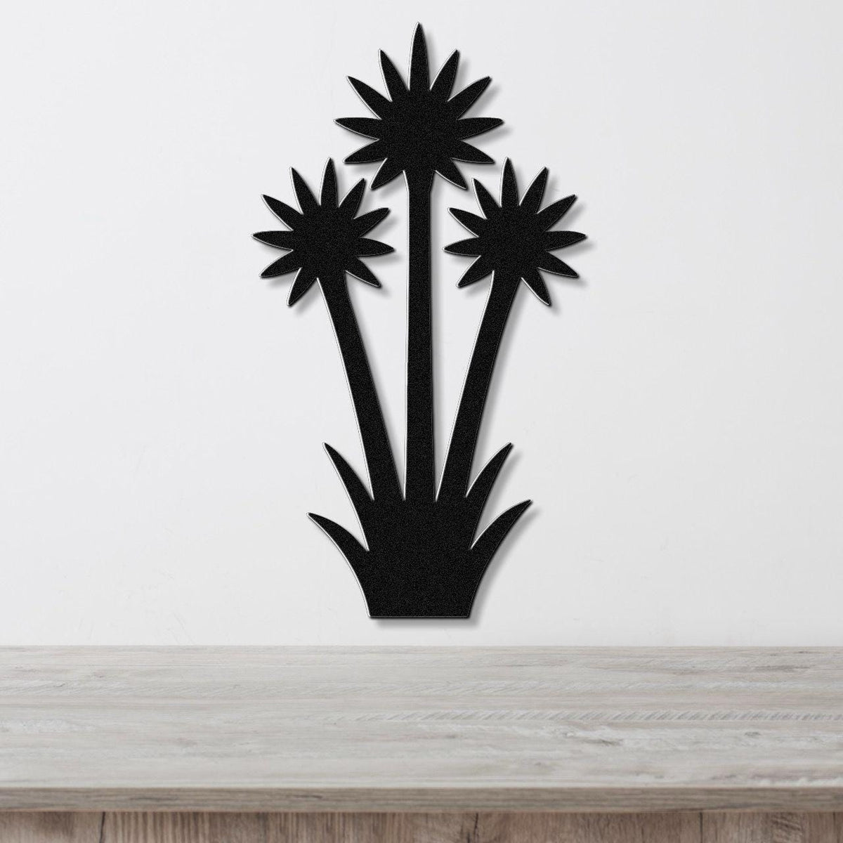 Black metal wall art cutout of three yucca plants with starburst tops, mounted on a white wall above a light wood surface.