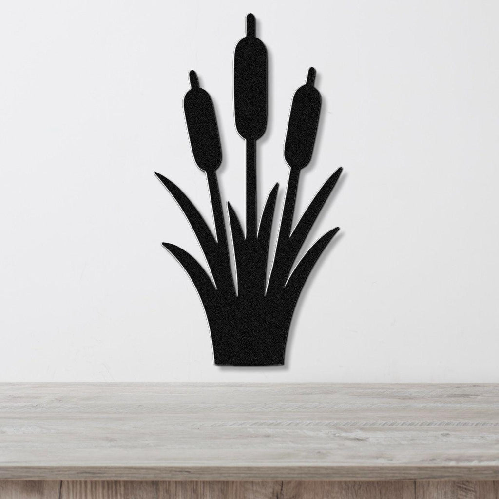 Black metal cattail wall art with three seed heads and leaf cutouts, mounted on a white wall above a light wood surface.