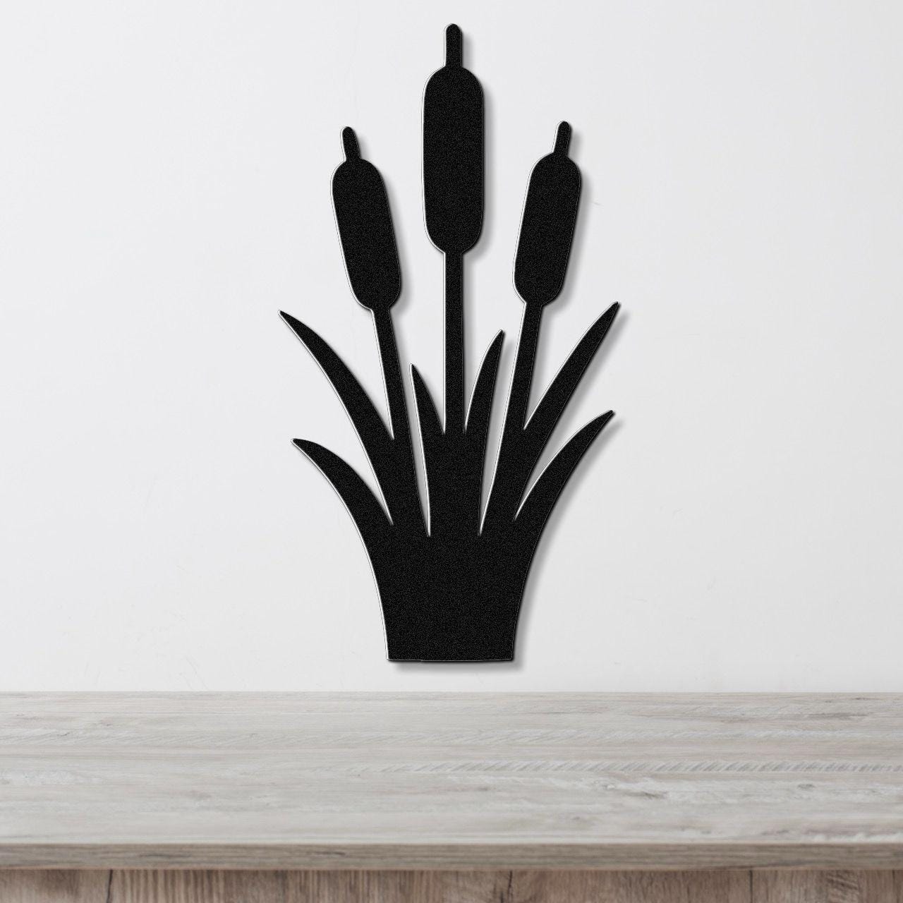 Black metal cattail wall art with three seed heads and leaf cutouts, mounted on a white wall above a light wood surface.