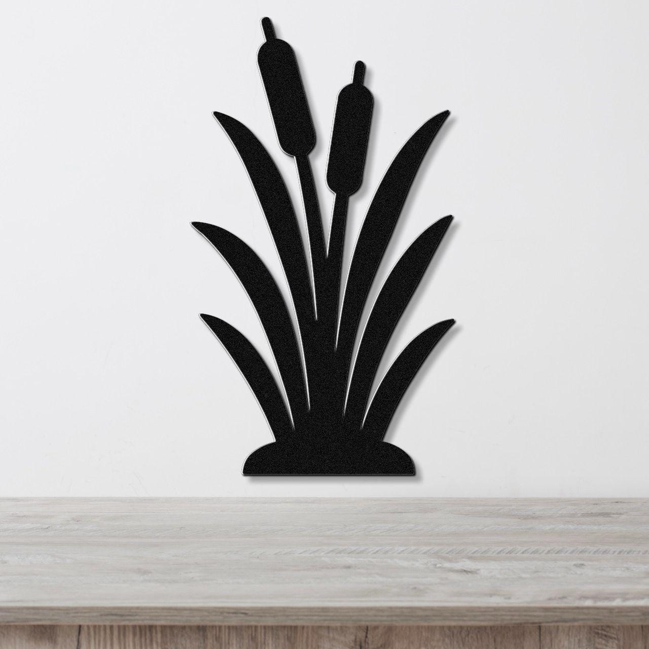 Black metal wall art in the silhouette of a cattail plant with two seed heads and long leaves, mounted on a white wall above a wooden surface.