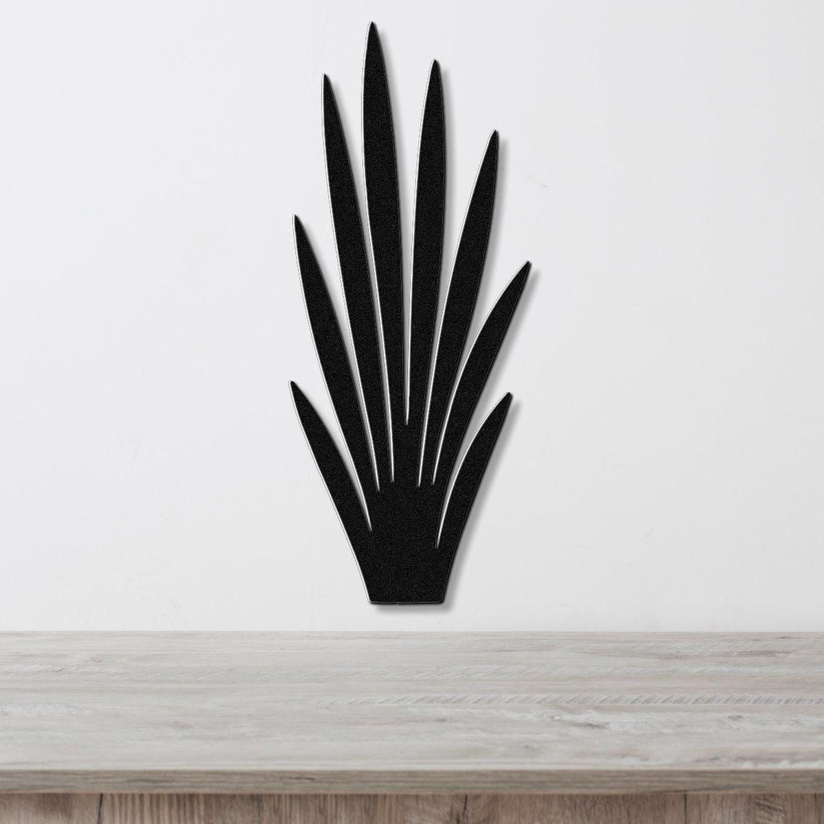 Black metal wall art in a silhouette of wild grass with pointed leaves, mounted on a white wall above a light wood surface.