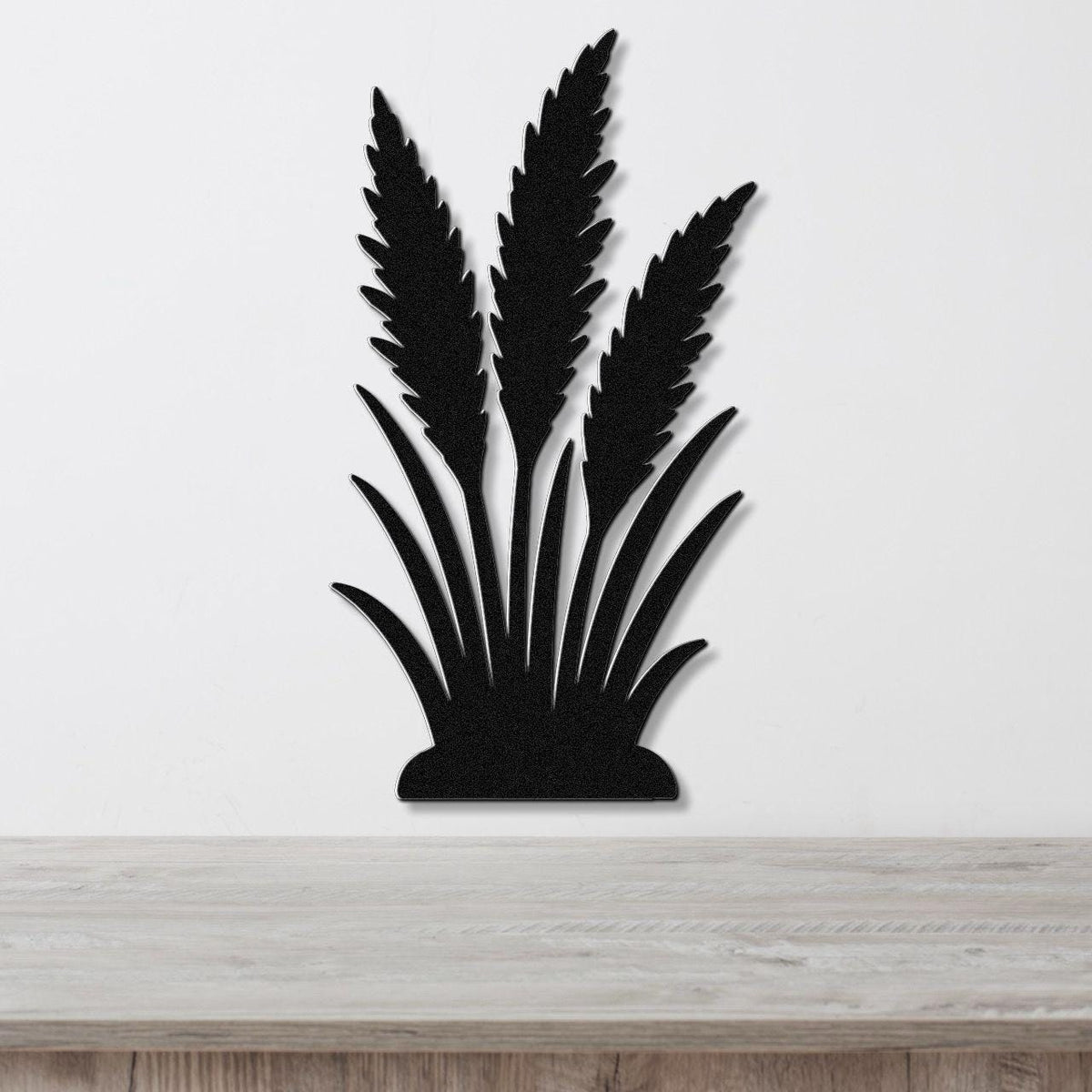 Black metal wall art in a silhouette of wild grass with layered blades and seed heads, mounted on a white wall above a light wood surface.