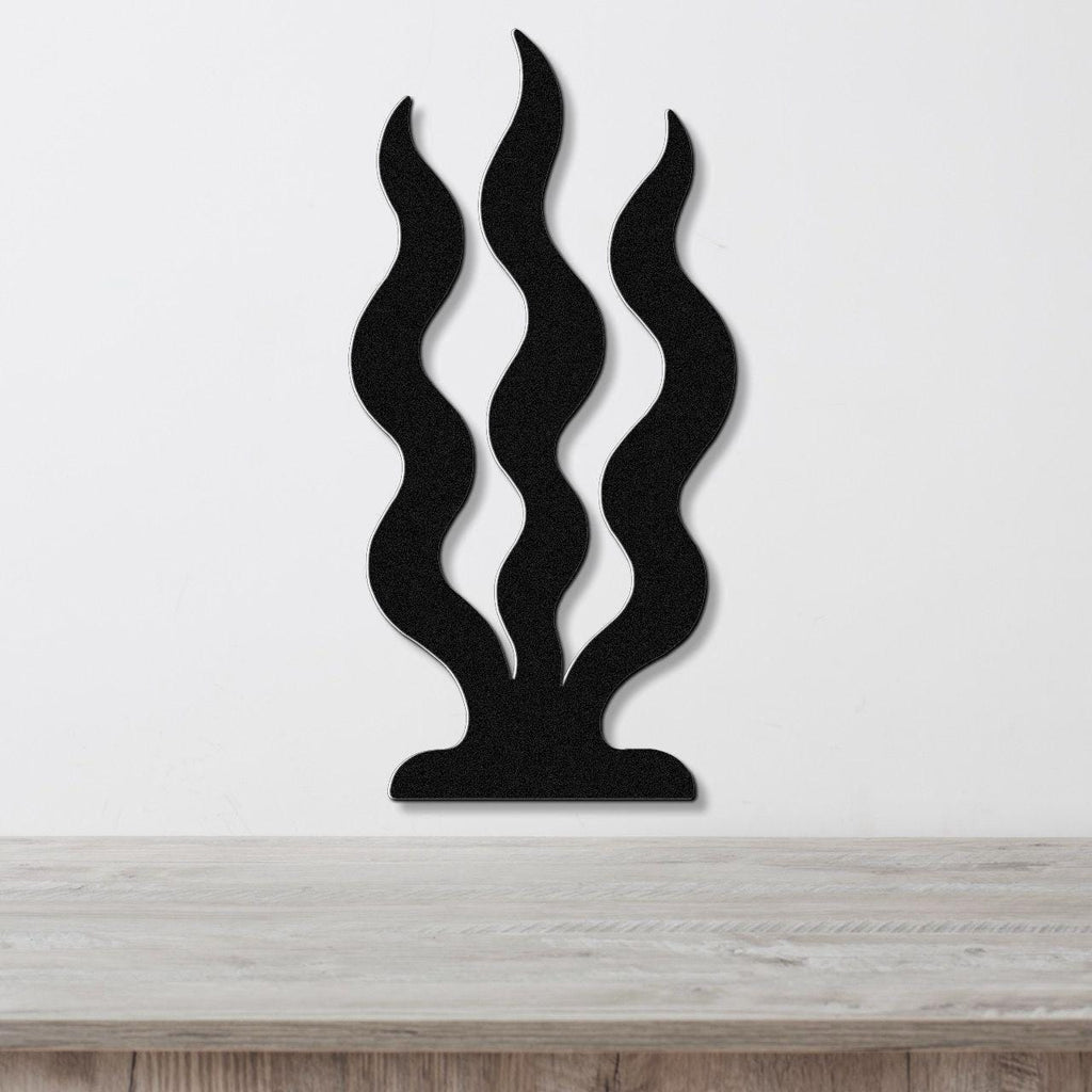 Black metal wall art with three wavy vertical cutouts resembling grass or seaweed, mounted on a white wall above a light wood surface.