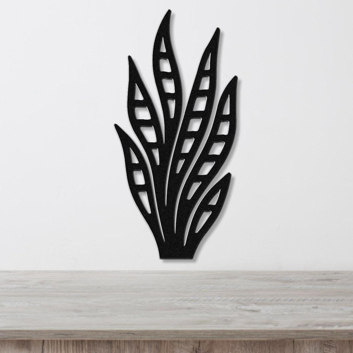 Black metal wall art in a snake plant silhouette with internal cutout details, mounted on a white wall above a light wood surface.