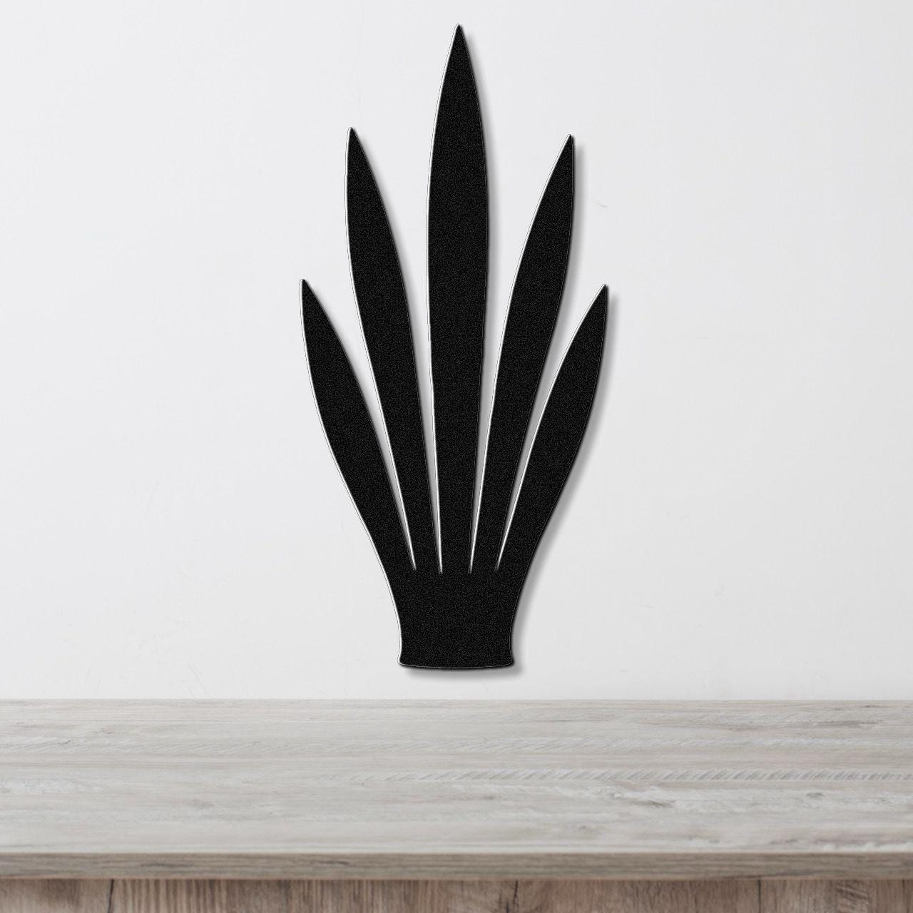 Black metal wall art in the shape of a snake plant with six pointed leaves, mounted on a white wall above a light wood surface.
