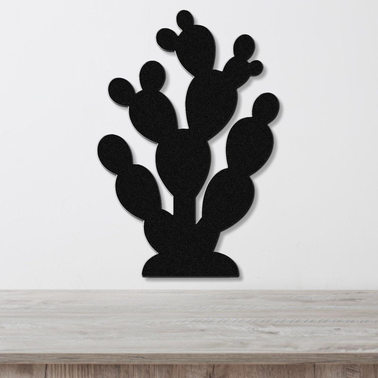 Black metal prickly pear cactus wall art with layered pads and cutout details, mounted on a white wall above a light wood surface.