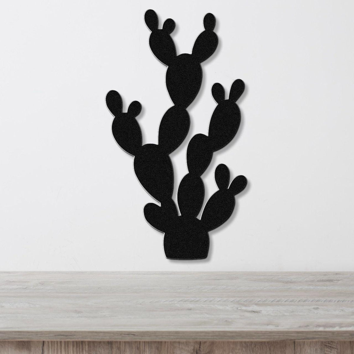 Black metal cactus wall art with multiple paddle-shaped segments, mounted on a white wall above a light wood surface.