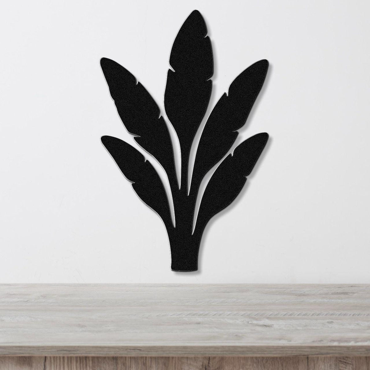 Black metal wall art in a banana plant silhouette with layered leaf cutouts, mounted on a white wall above a light wood surface.