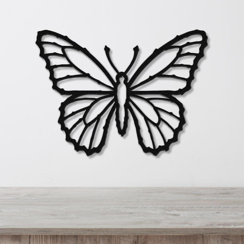 Black metal butterfly wall art with detailed wing cutouts, mounted on a white wall above a light wood surface.