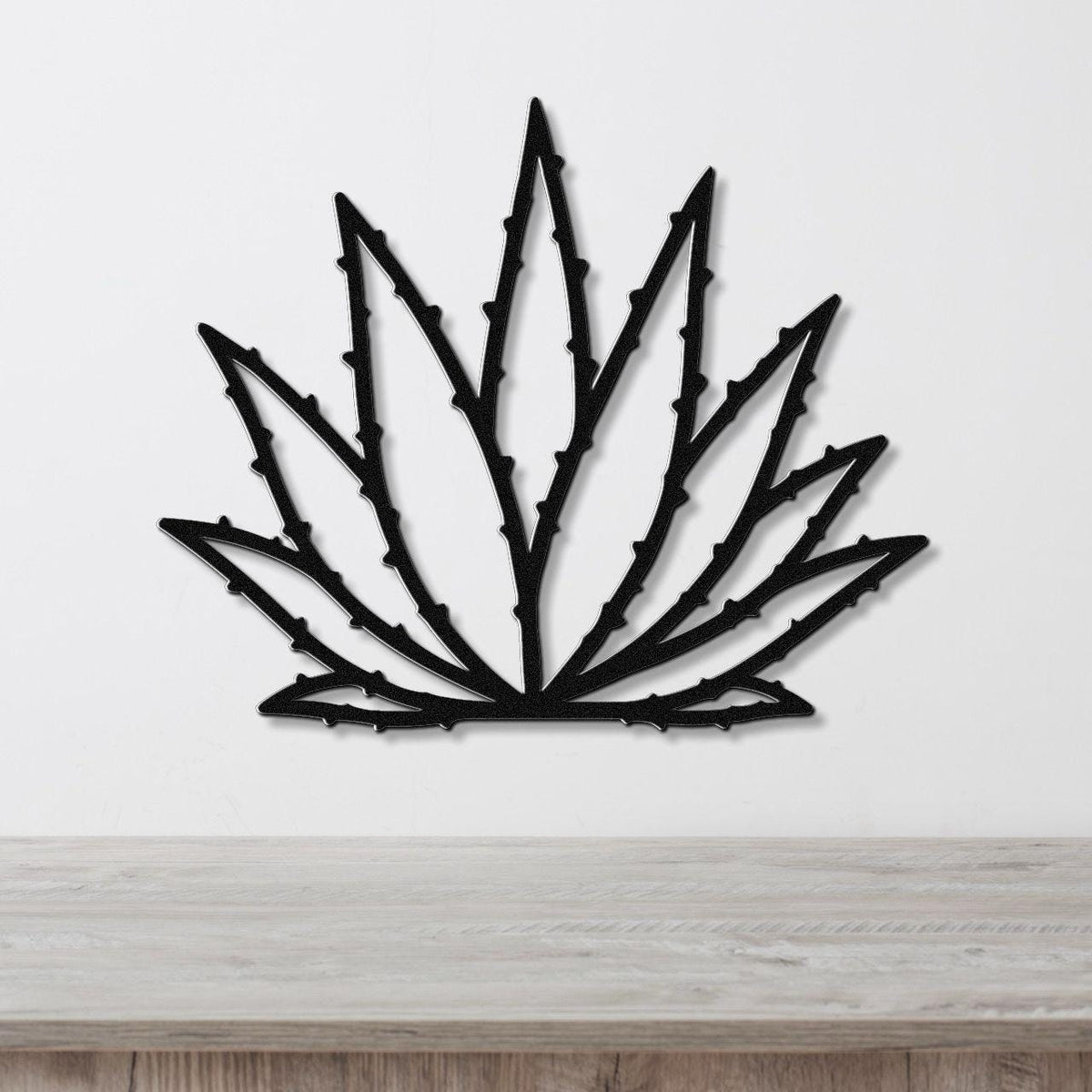 Black metal agave wall art with spiky leaf cutouts, mounted on a white wall above a light wood surface.