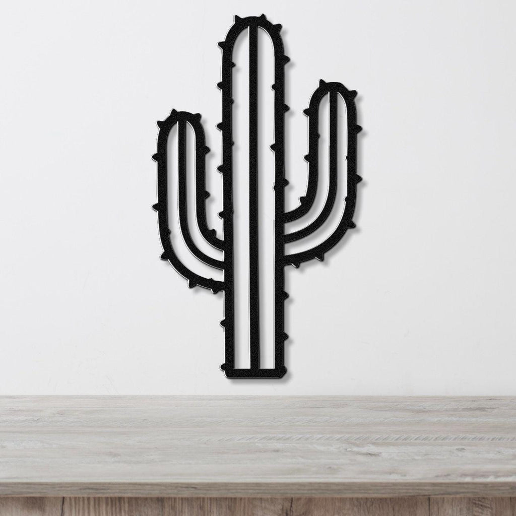 Black metal wall art in the silhouette of a saguaro cactus with spines and side arms, mounted on a white wall above a light wood surface.
